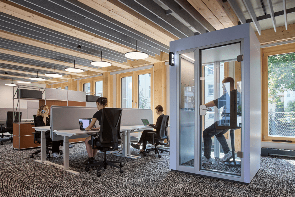Modern office with people working at desks and one person inside a glass phone booth, surrounded by wooden beams and windows.