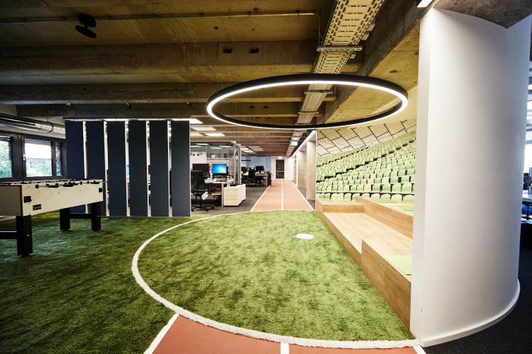 An office with green turf, a foosball table, stadium seating, and a circular light fixture above.