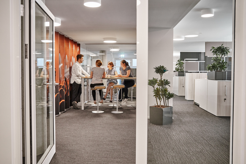 Four people sit at a high table in a modern office with glass walls, plants, and white cabinets.