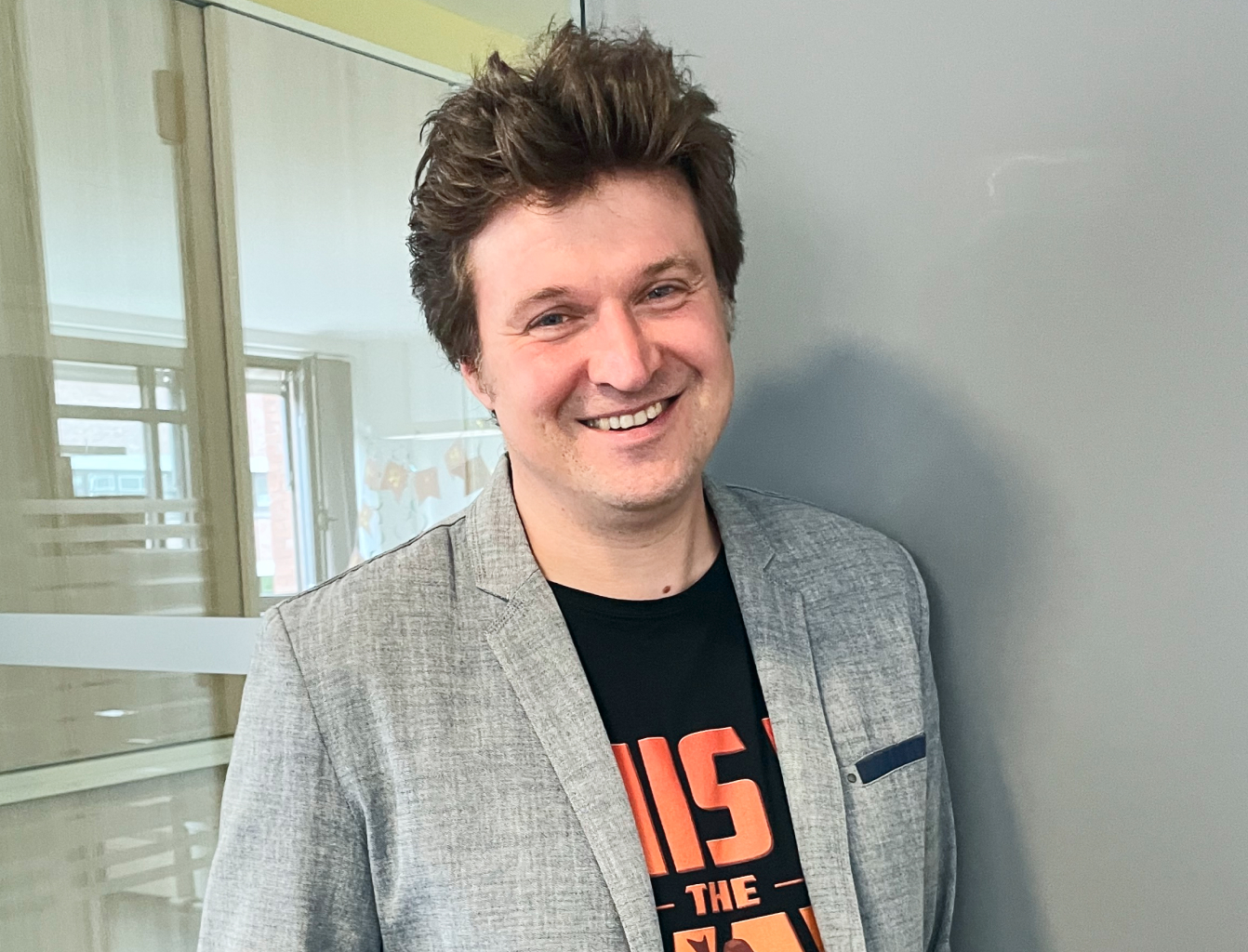 Smiling man in a gray blazer and black t-shirt stands indoors against a gray wall, with windows in the background.