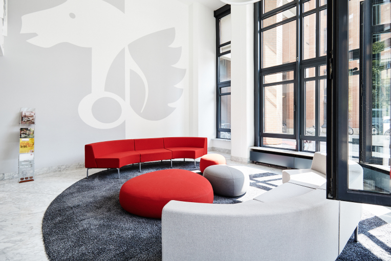 Modern lobby with red and gray curved sofas, round ottomans, large windows, and a wall mural of a winged creature.