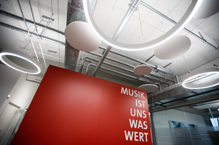 Red wall with German text, circular ceiling lights, and visible industrial pipes in a modern interior space.