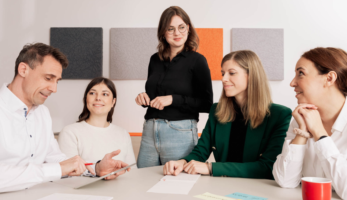 Five people in a meeting, three seated, two standing, with papers and a red mug on the table. Colorful panels on the wall.