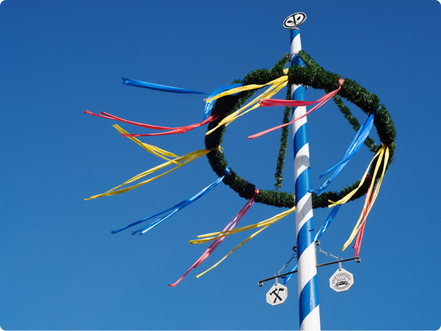 Maibaum mit grünem Kranz und bunten Bändern vor blauem Himmel.
