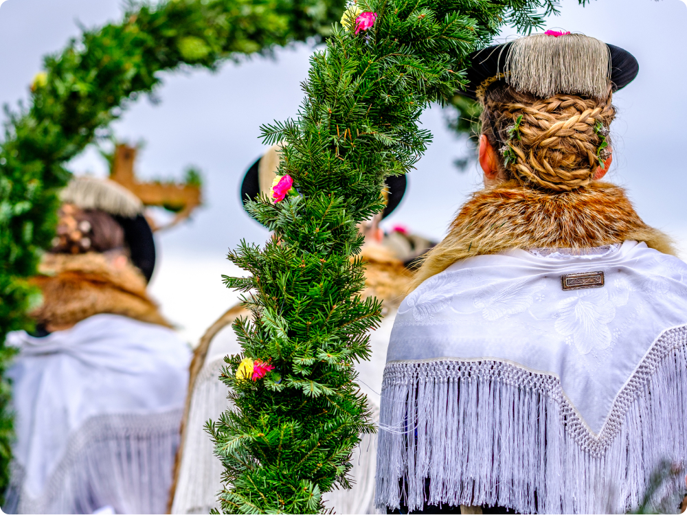 Rückansicht von Personen in traditioneller Tracht mit geflochtenem Haar, Pelzkragen und weißen Fransen, davor ein geschmückter grüner Kranz.