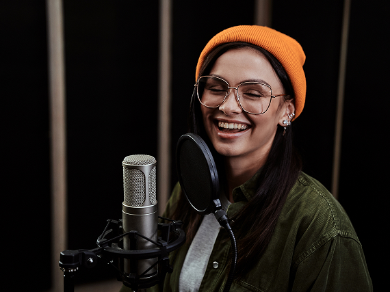 Eine lächelnde Person mit Brille und orangener Mütze singt in ein Mikrofon im Studio.