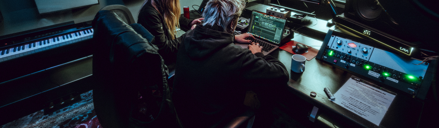 Zwei Personen arbeiten an einem Laptop in einem Musikstudio mit Keyboard und Mischpult.