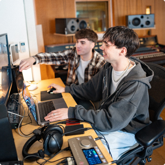 Zwei Personen arbeiten lächelnd an Computern in einem Tonstudio mit Lautsprechern und Kopfhörern.