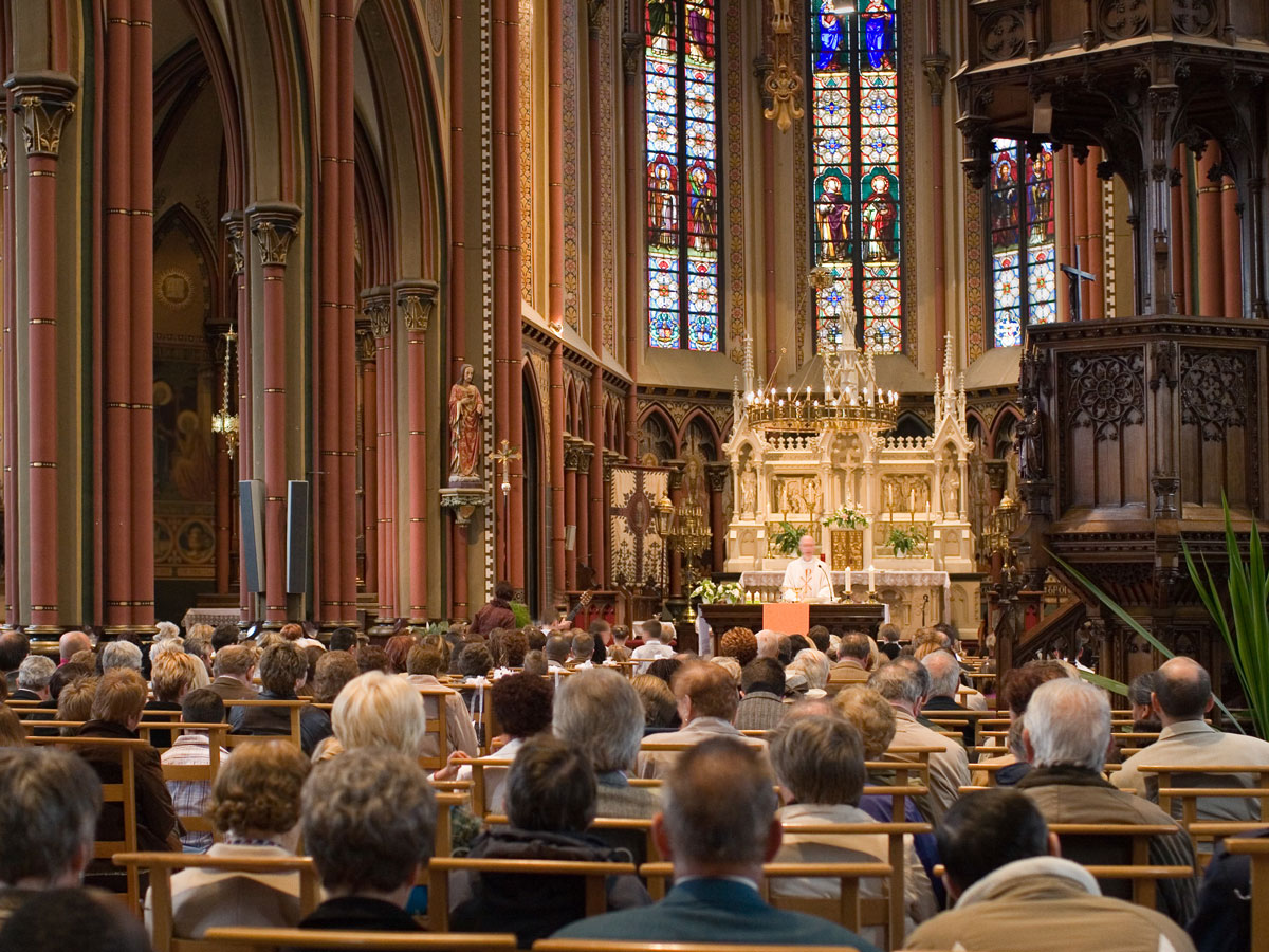 A priest speaks at an ornate church altar with stained glass windows, facing a seated congregation.