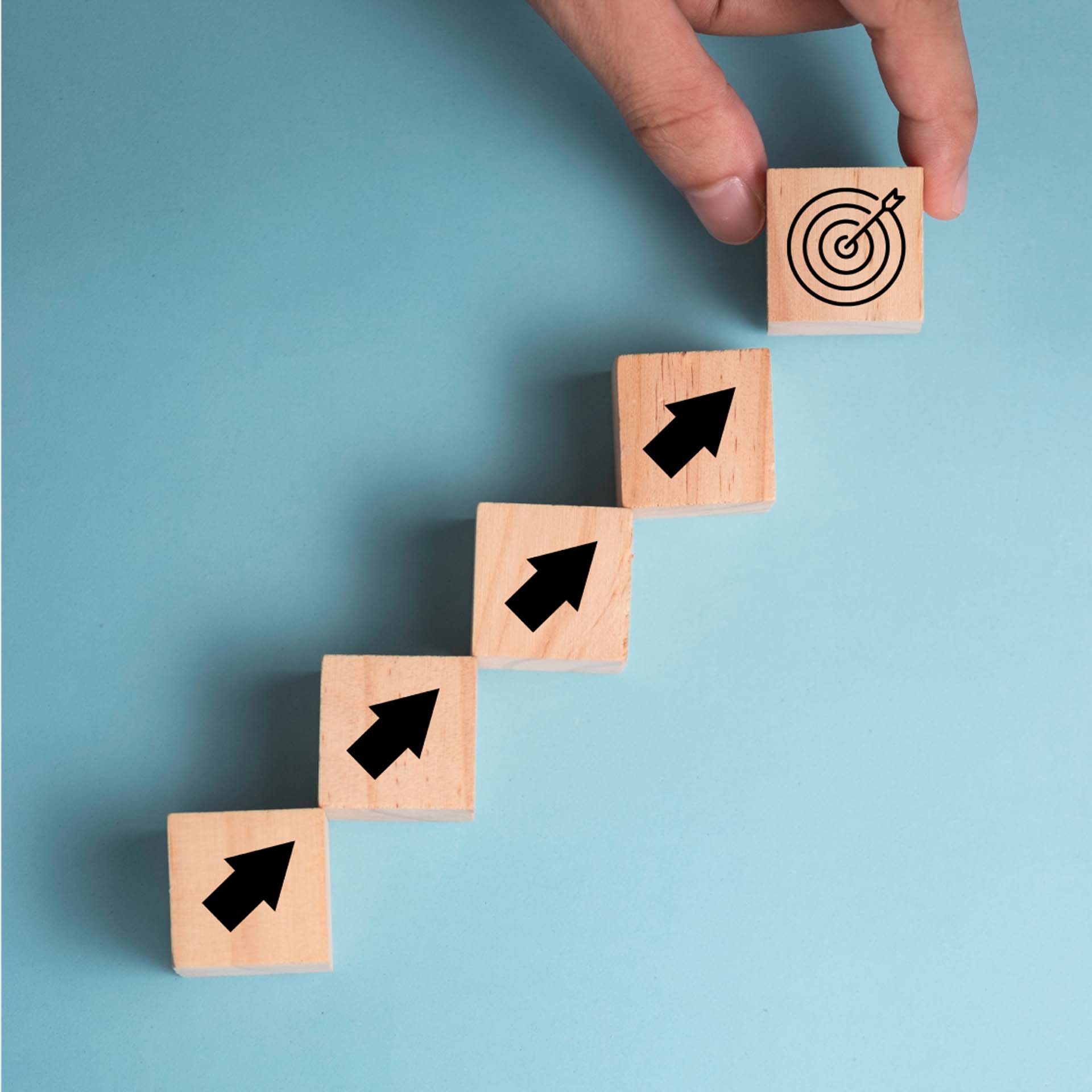 A hand arranges wooden blocks in a staircase pattern, each with an arrow, ending in a target symbol on a blue background.
