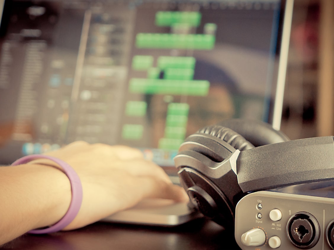 A hand with a purple bracelet on a laptop keyboard, headphones, and audio equipment nearby, with a blurred screen in the background.