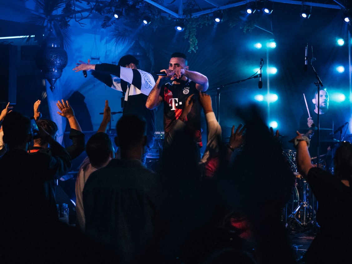 A musician performs on stage with a drummer, surrounded by blue lights and a cheering crowd.
