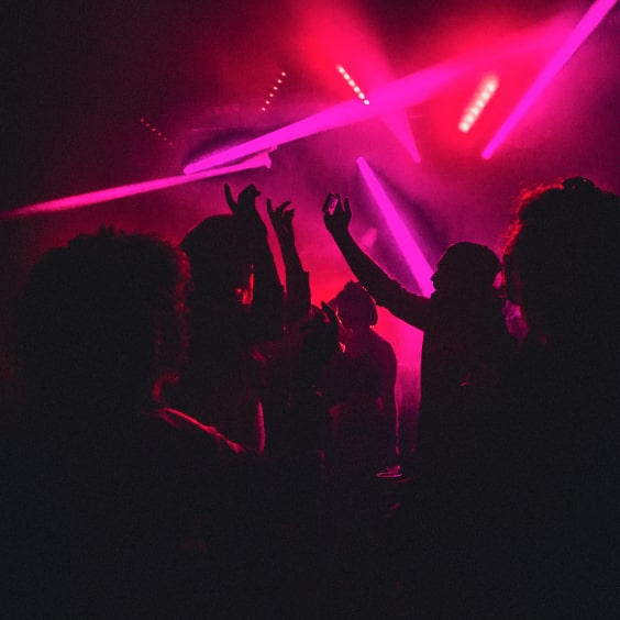 Silhouetted people dance under vibrant pink and purple lights in a dark club setting.