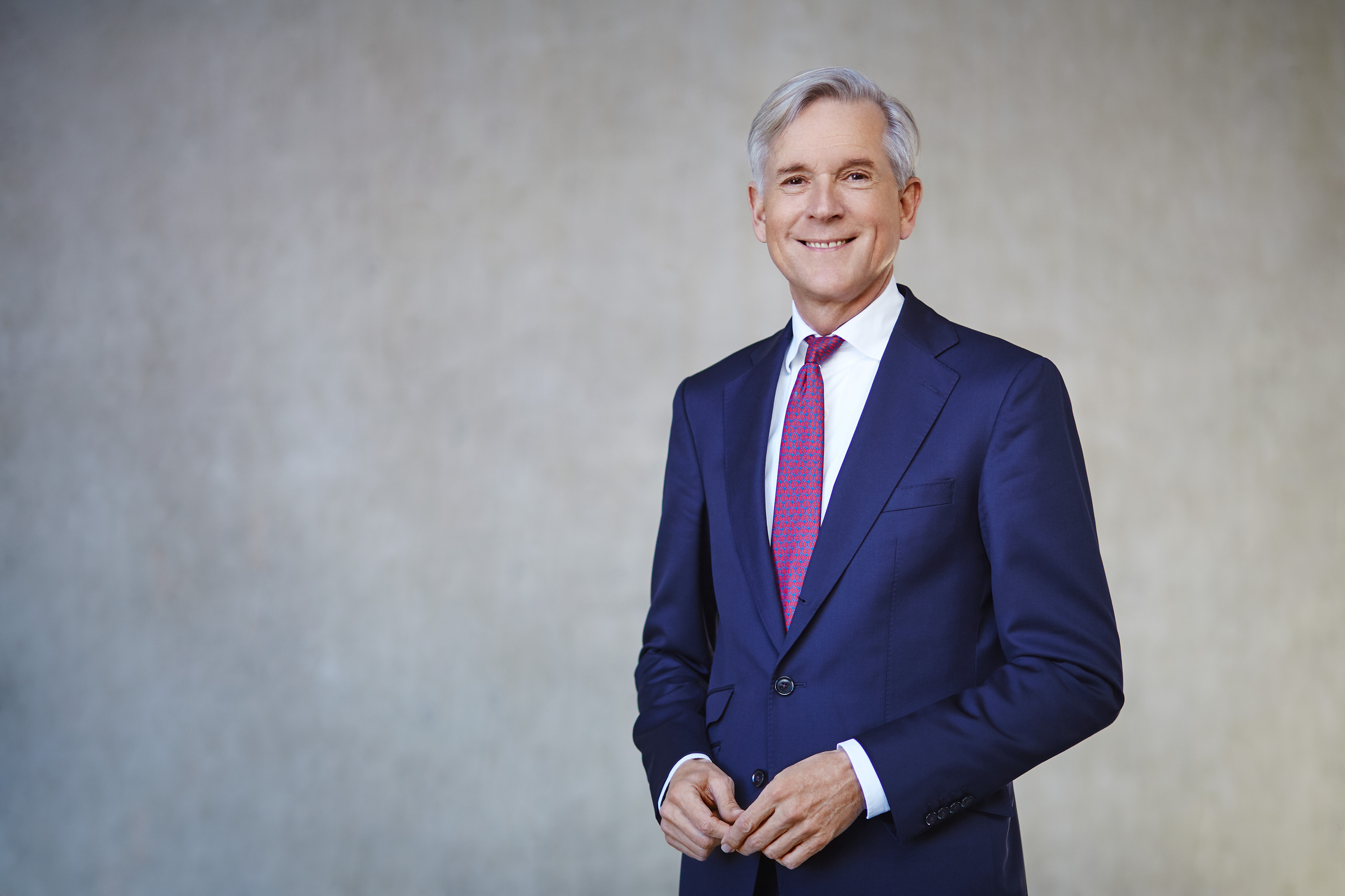 Smiling man in a navy suit with a red tie stands against a light gray background.