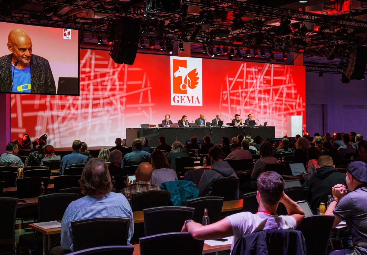 A conference with a panel of six people on stage, red GEMA logo backdrop, and audience seated facing them.