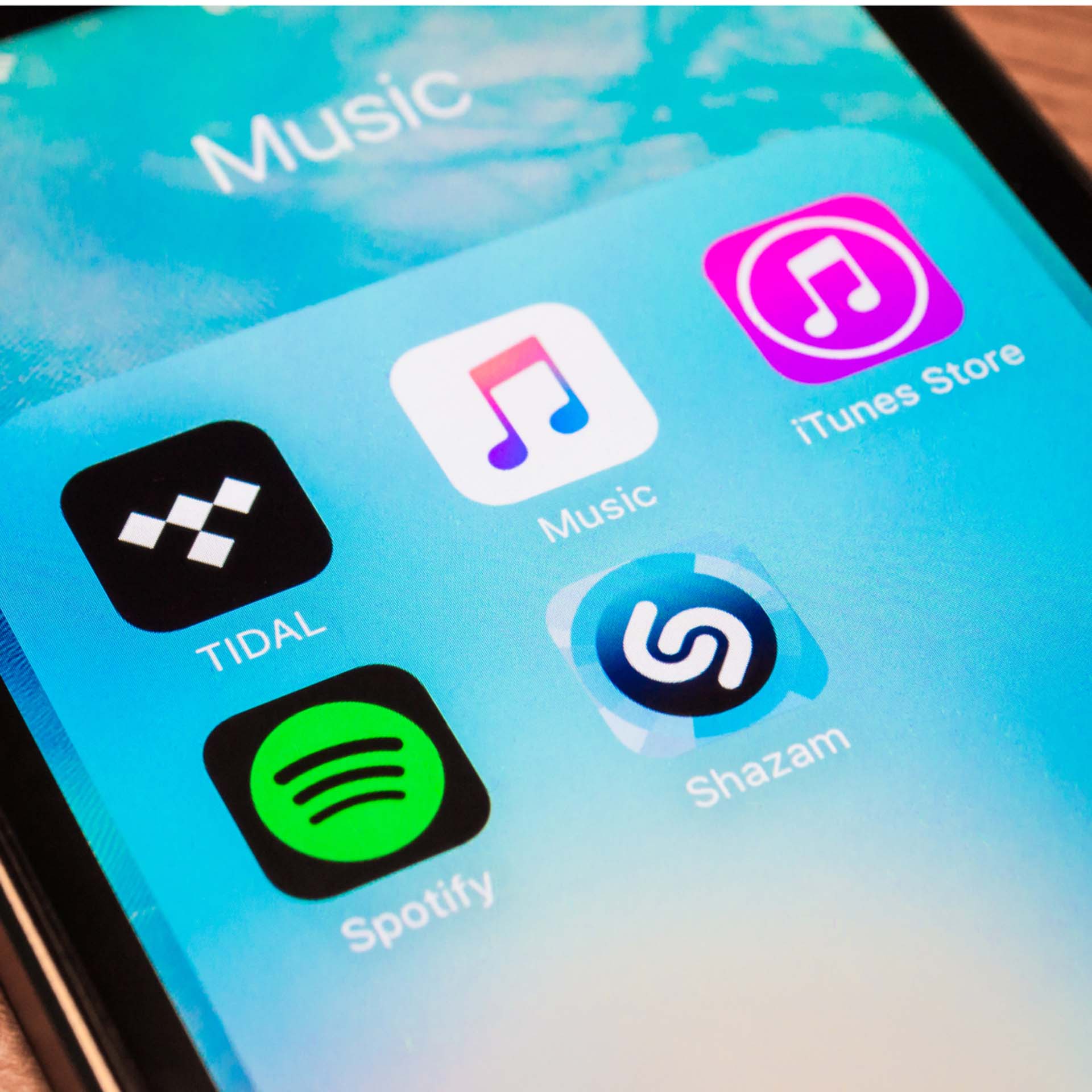 Smartphone screen showing music apps: Tidal, Music, iTunes Store, Spotify, and Shazam on a blue background.