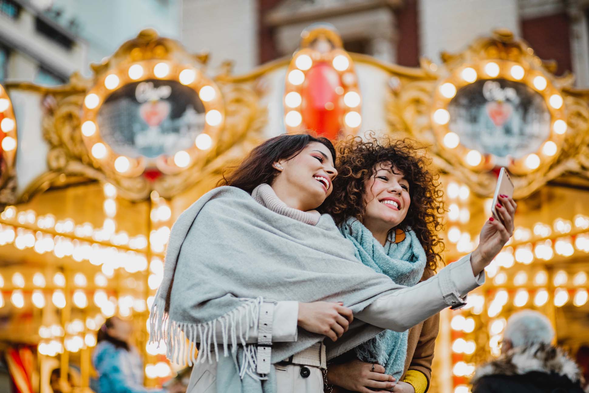 Zwei lachende Frauen machen ein Selfie vor einem beleuchteten Karussell.
