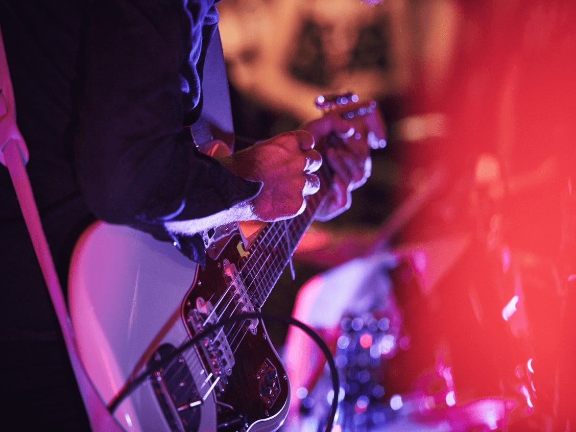 A person plays an electric guitar under red and purple stage lights.