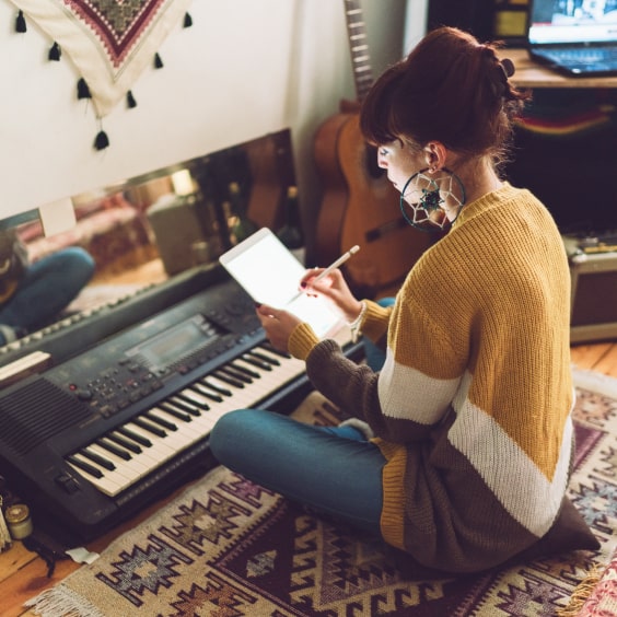 A person in a cozy sweater writes on a tablet beside a keyboard, sitting on a patterned rug with a guitar nearby.