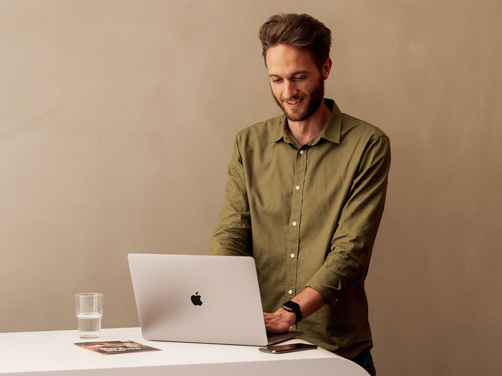 Man in a green shirt uses a laptop on a white table with a glass of water and a magazine nearby.