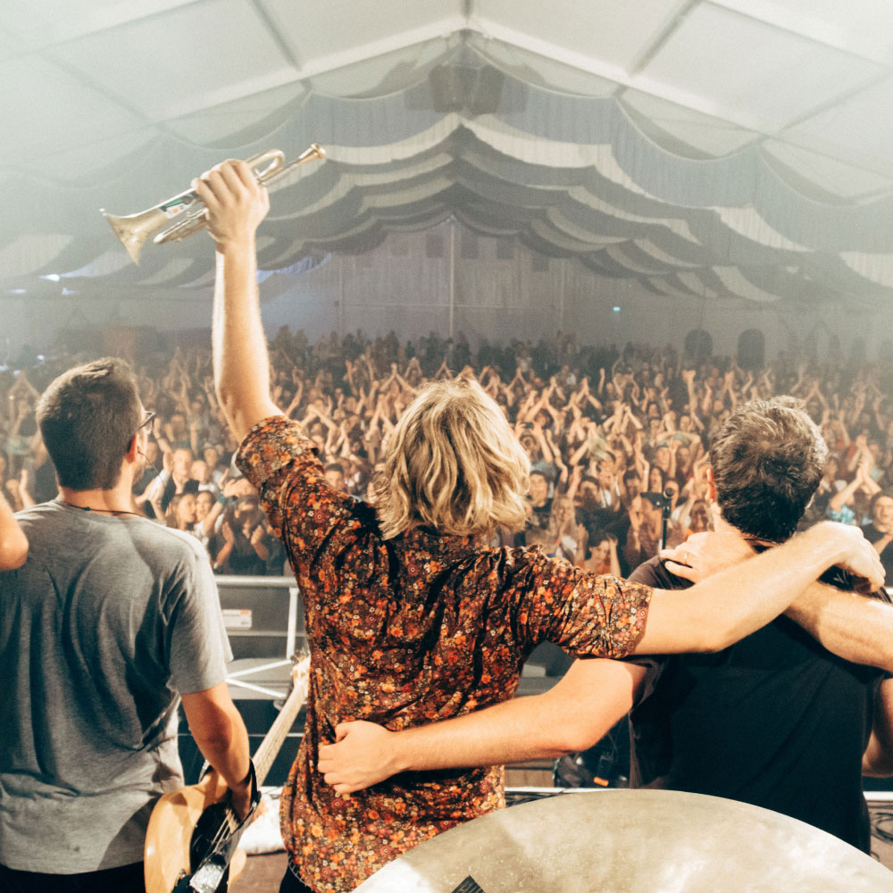 Three musicians on stage face a cheering crowd, one holding a trumpet aloft, in a large tent with draped ceiling fabric.