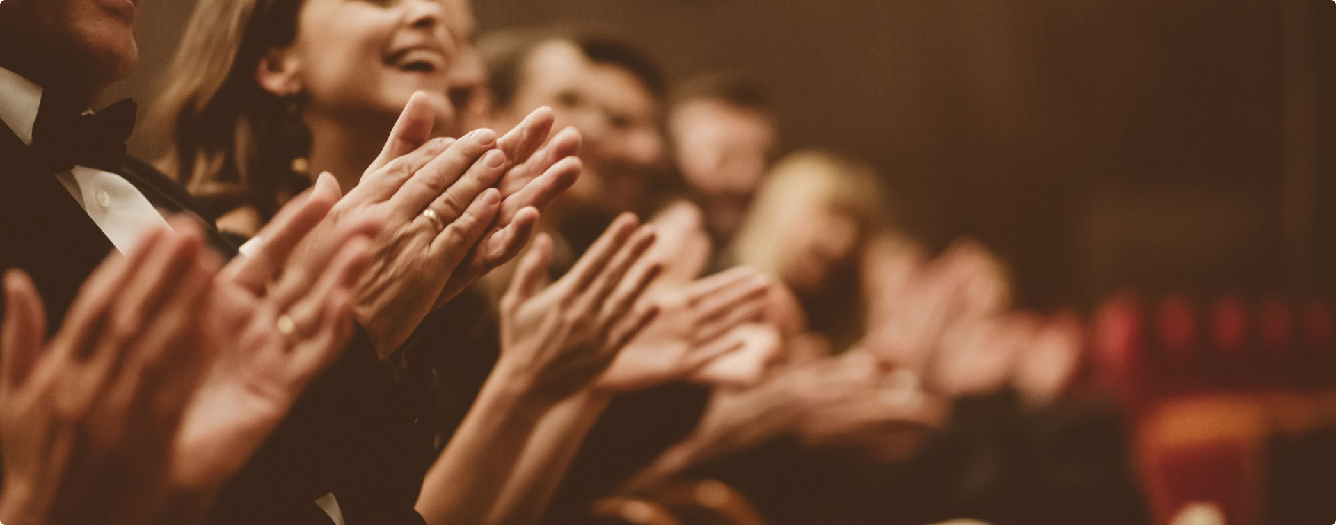 People in formal attire clapping enthusiastically in a dimly lit setting.