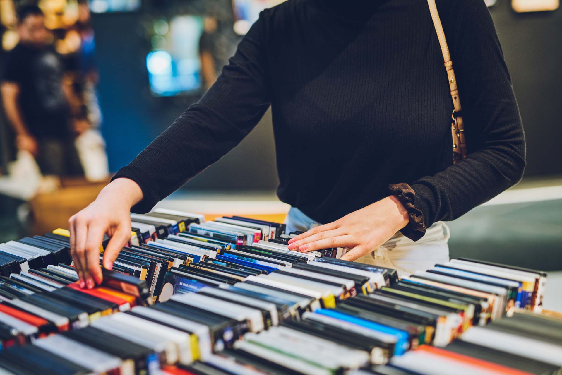 Person in schwarzem Pullover stöbert durch eine Sammlung von DVDs oder Büchern auf einem Tisch.