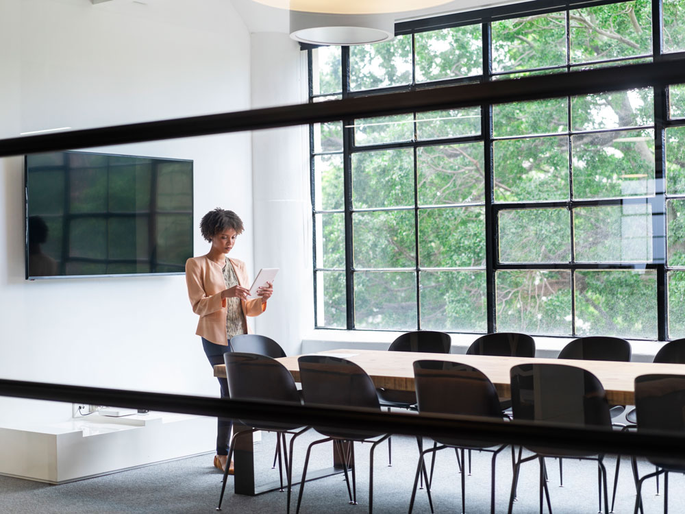 Frau mit Tablet steht in modernem Konferenzraum, große Fenster im Hintergrund.