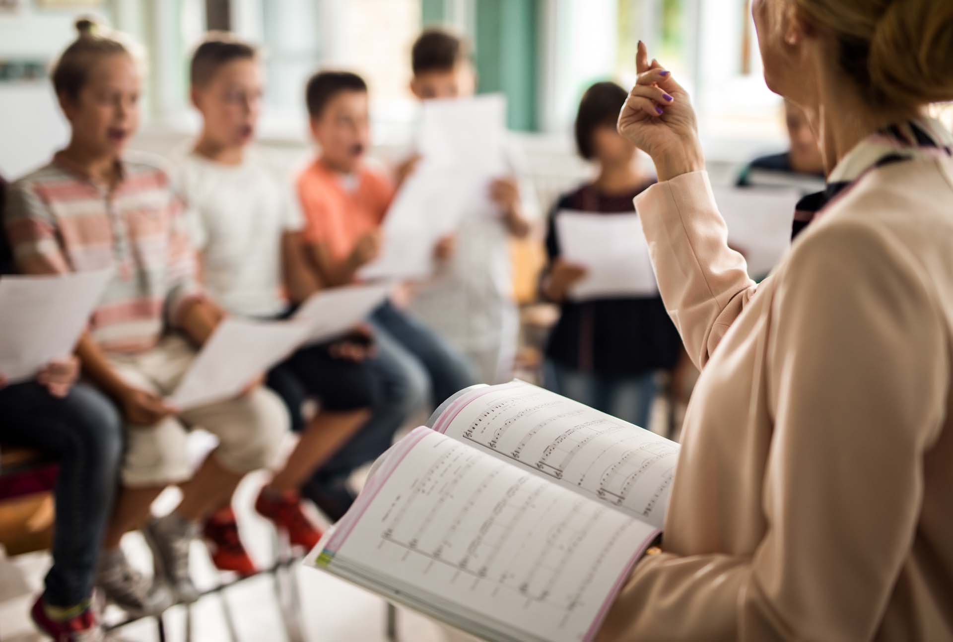 Eine Lehrerin dirigiert singende Kinder, die Notenblätter halten.