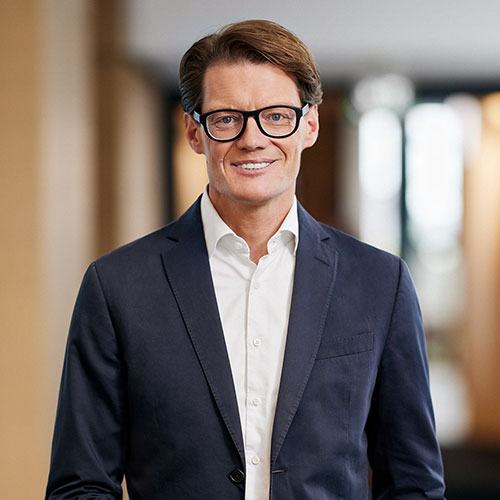 A person in a dark suit and glasses smiles, standing indoors with a blurred background.