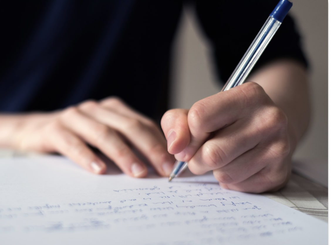 A person writes with a blue pen on paper, with handwritten text visible.