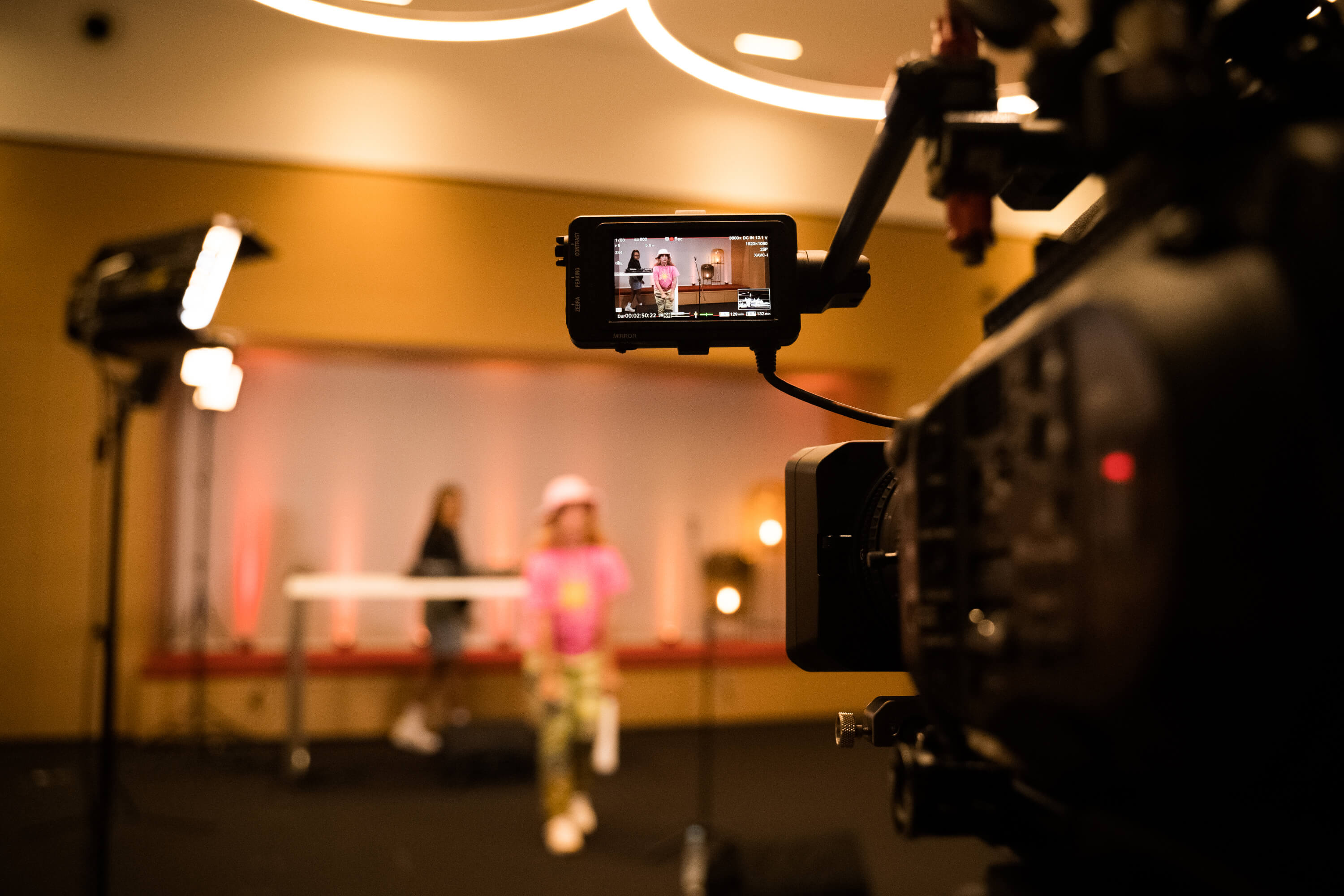 A video camera films a person in pink and a DJ in the background, with studio lights and a blurred setting.
