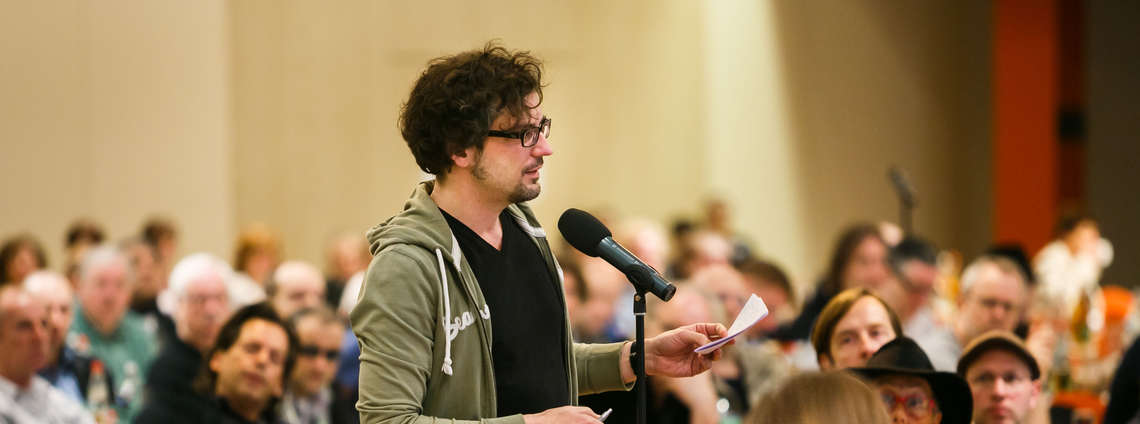 A man in a green hoodie speaks into a microphone in a crowded room, holding a piece of paper.
