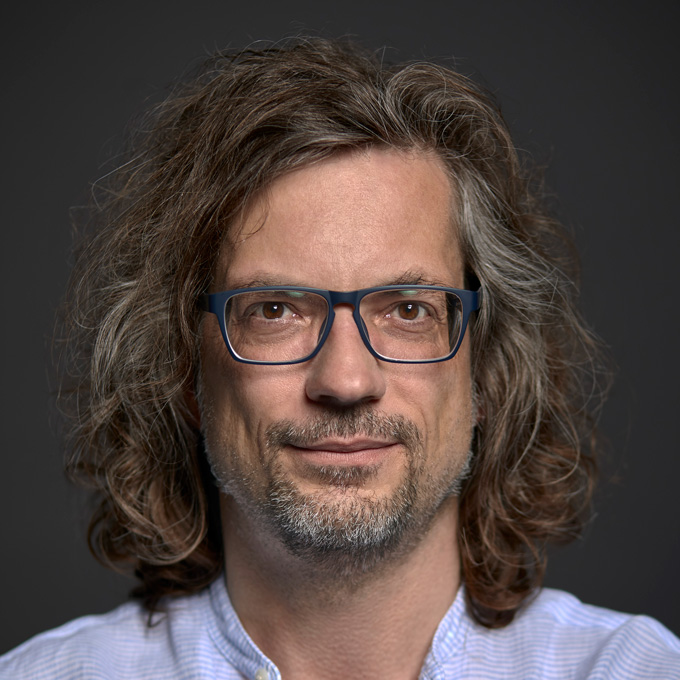 Eine Person mit langer, lockiger Frisur und Brille vor grauem Hintergrund.