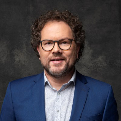 Ein Mann mit Brille in blauem Anzug vor dunklem Hintergrund.