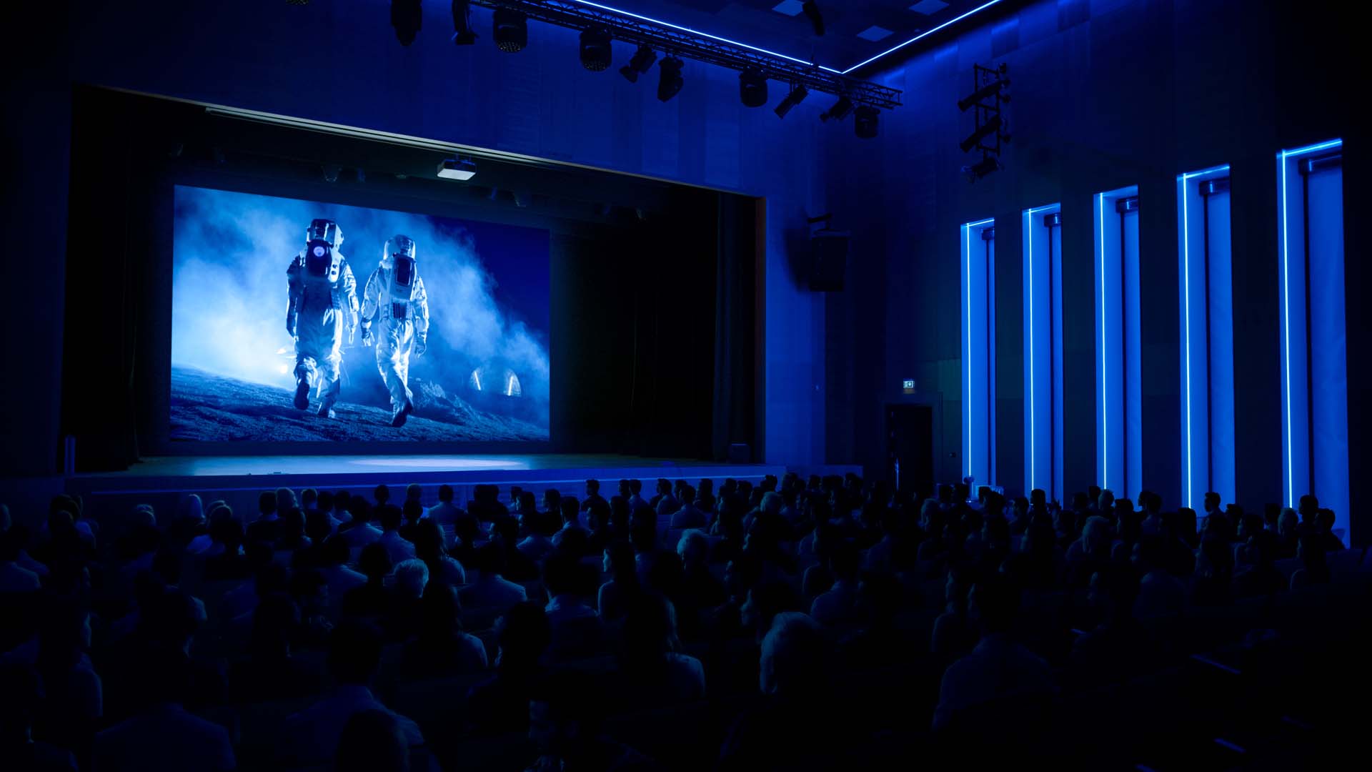 A dimly lit theater with a blue glow shows a large screen displaying two astronauts walking on a misty surface.
