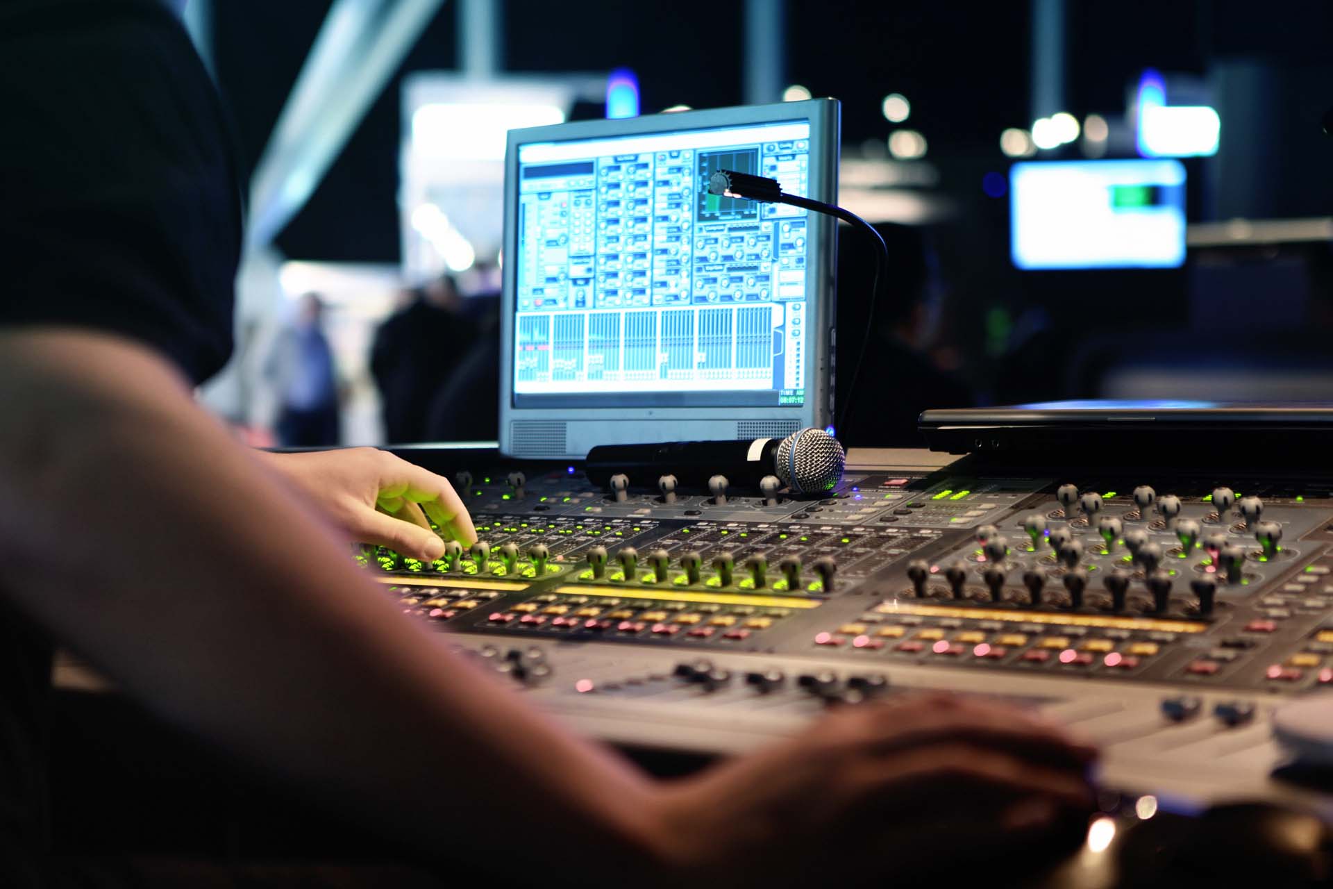 Hands adjust audio mixer with glowing controls, next to a computer screen displaying sound levels, and a microphone.