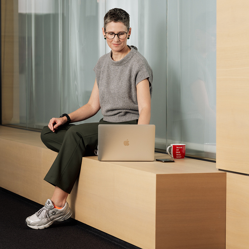 Person in gray top and green pants sits on a bench with a laptop, red mug, and phone nearby.