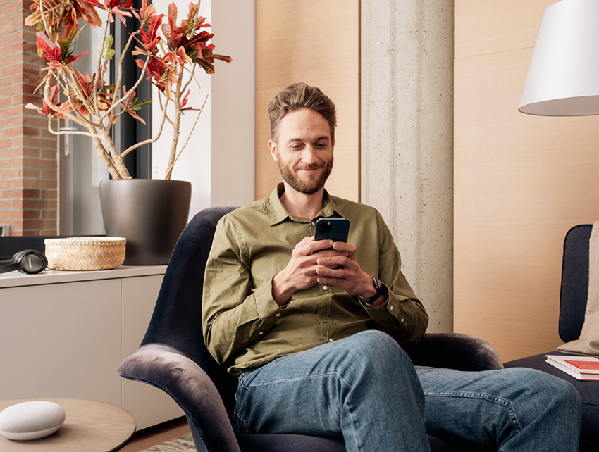 A man in a green shirt sits in a chair, smiling at his phone. A plant and lamp are in the background.
