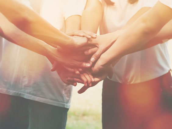 A group of people stack their hands together in a show of unity, with soft sunlight filtering through.