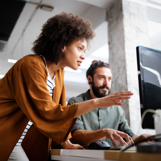 Two people collaborate at a computer, one pointing at the screen, in a modern office setting.