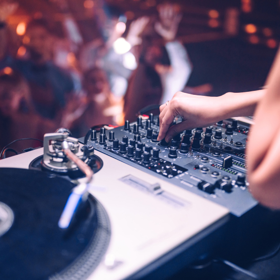 A DJ adjusts a mixer next to a turntable, with a crowd dancing in the blurred background.