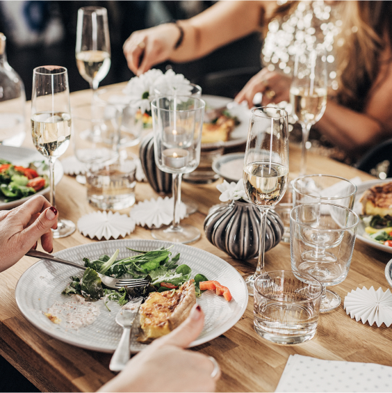 Ein gedeckter Tisch mit Salat, Quiche und Sektgläsern, umgeben von Menschen in festlicher Atmosphäre.
