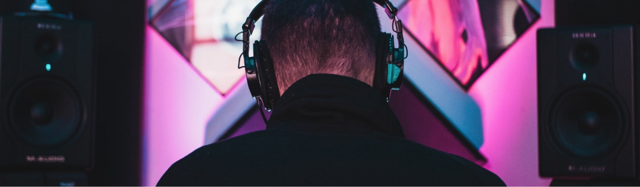 Person wearing headphones facing speakers, with a pink and blue lit background.