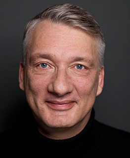 Smiling man with short gray hair, wearing a black turtleneck, against a dark background.
