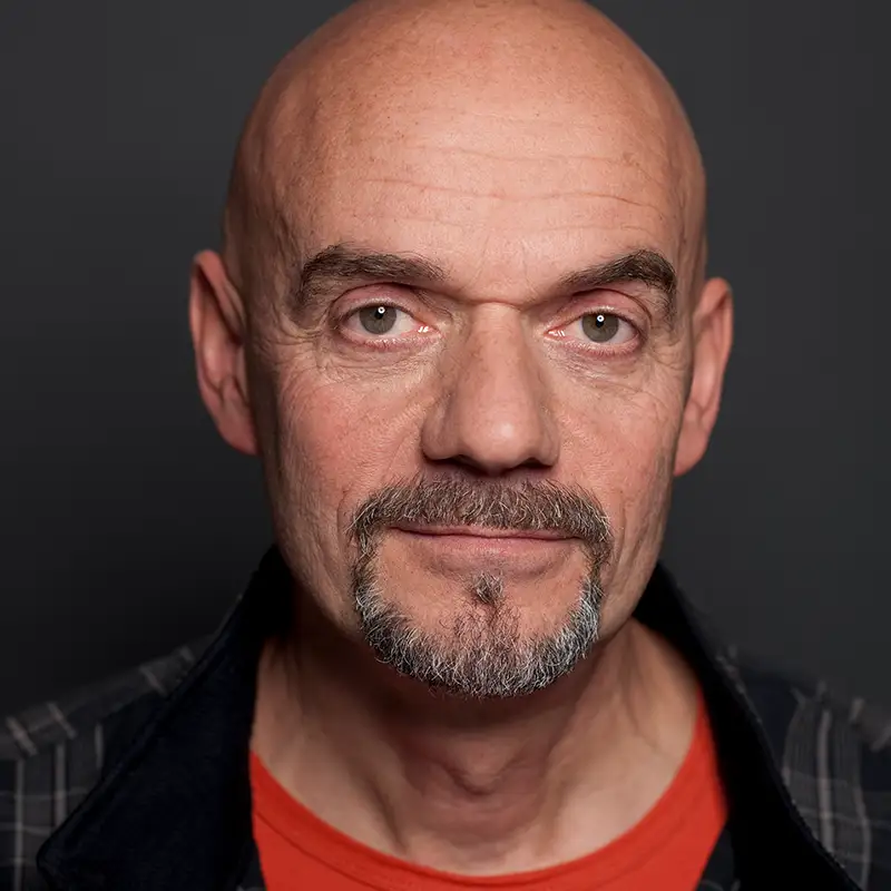 Ein Mann mit Glatze und Bart trägt ein rotes Hemd vor dunklem Hintergrund.