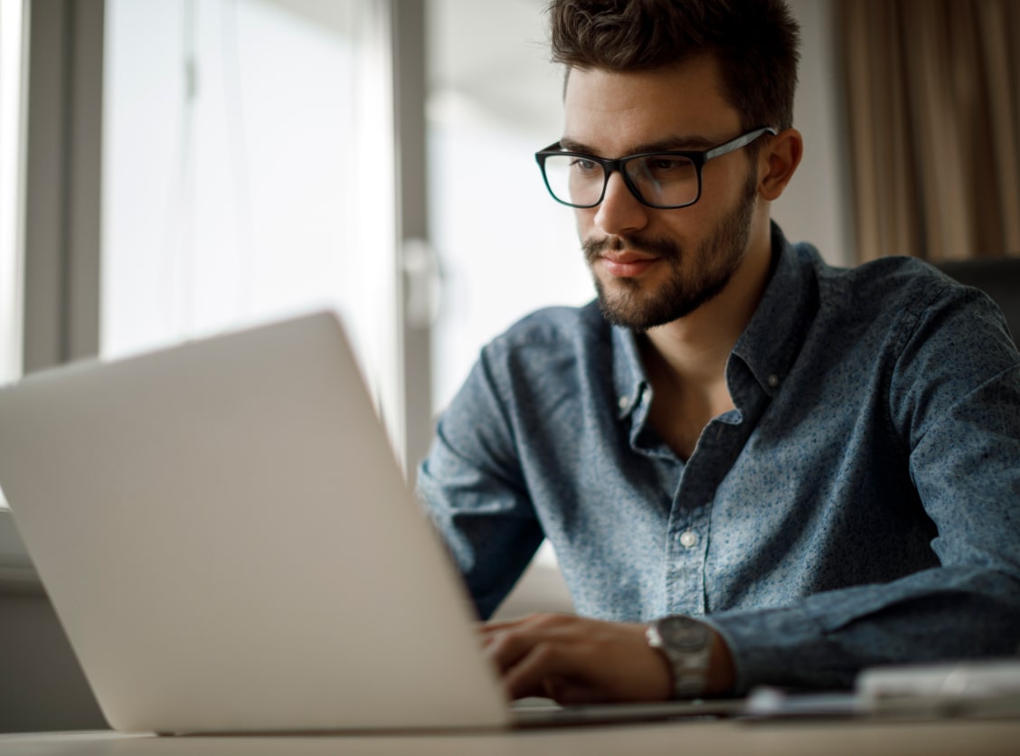 A man with glasses works on a laptop in a bright room.