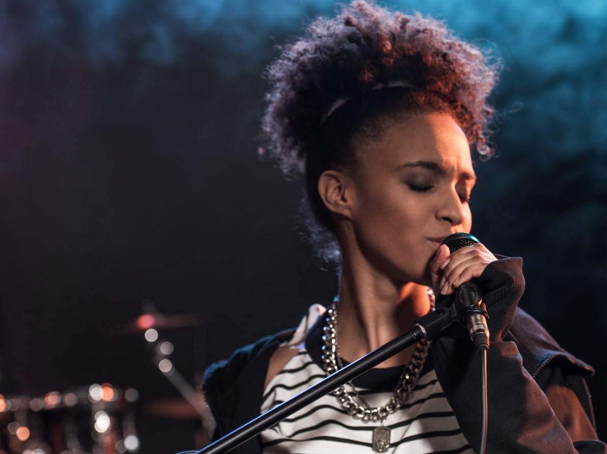 Singer with curly hair and striped shirt passionately sings into a microphone on stage with drums blurred in the background.