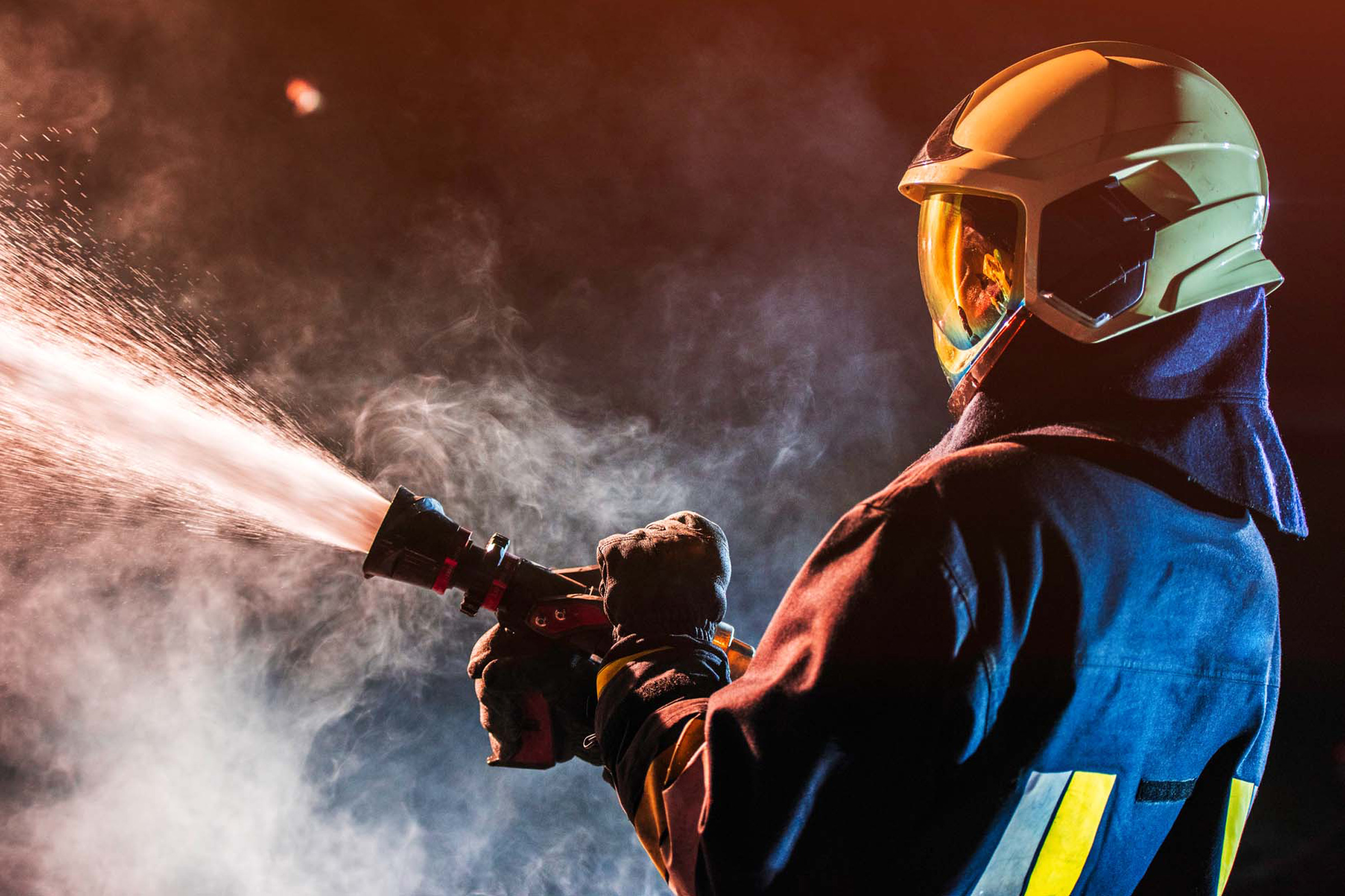 Ein Feuerwehrmann sprüht Wasser aus einem Schlauch, umgeben von Rauch und in Schutzkleidung mit Helm.