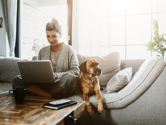 Woman using a laptop on a sofa with a dog beside her, a coffee cup and notebook on a wooden table in a bright room.